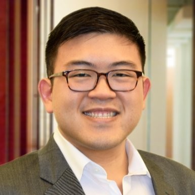 A close-up photo of a man with short black hair, wearing glasses and a gray suit jacket over a white collared shirt. He is smiling at the camera. The background is a blurred indoor setting with warm lighting, featuring modern office decor with red and beige tones.