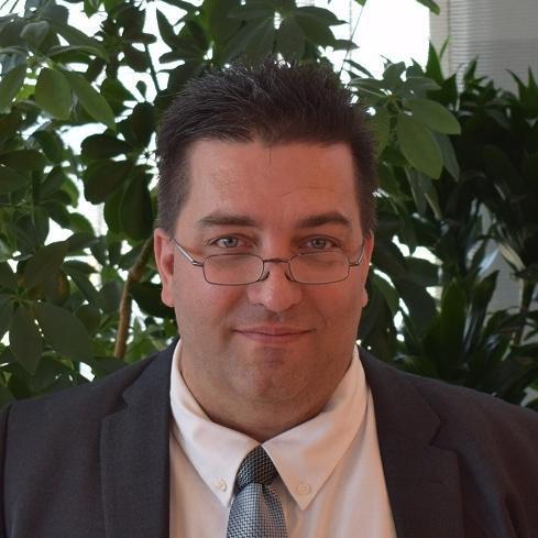 A close-up photo of a man with short dark hair, wearing glasses, a white dress shirt, a patterned tie, and a dark suit jacket. He is smiling slightly at the camera. The background shows green leafy plants, giving the image a natural setting.