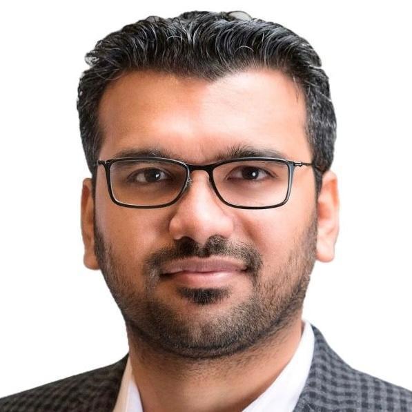 A close-up photo of a man with short black hair and a neatly trimmed beard, wearing glasses. He is dressed in a checkered suit jacket over a white shirt. The background is plain white, focusing attention on his face.