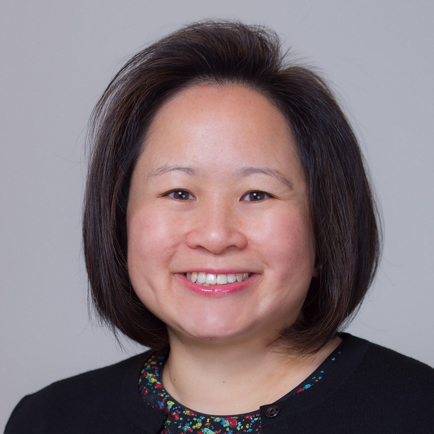 A woman smiles brightly at the camera. She has chin length brown hair and is wearing a black shirt. The background is grey.