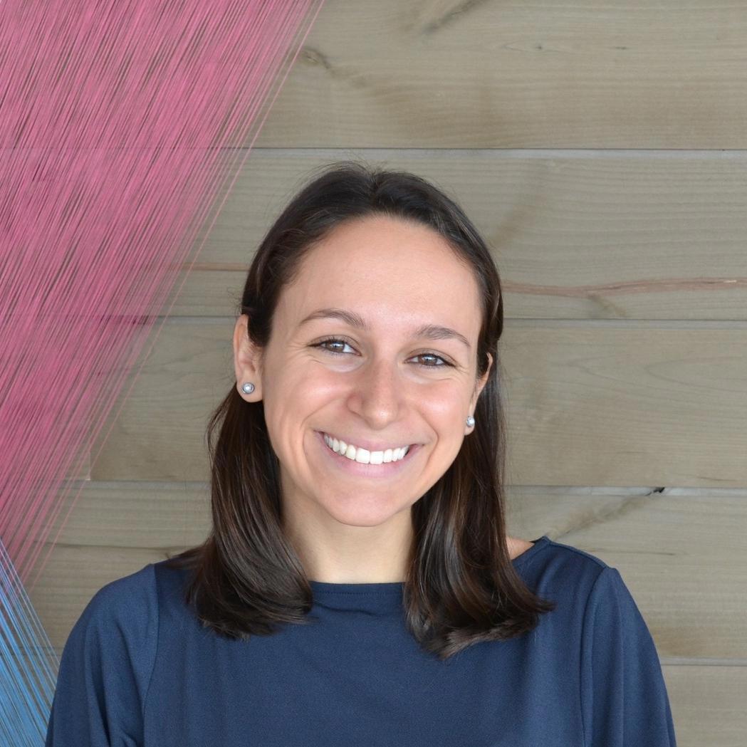 A close-up photo of a woman with shoulder-length brown hair, smiling brightly at the camera. She is wearing a dark blue top and small, round earrings. The background features a wooden panel wall with an artistic design of pink and blue strings arranged in a triangular pattern.