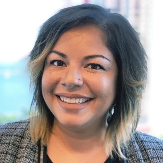 A close-up photo of a woman with shoulder-length dark hair that transitions to a lighter shade at the ends. She is smiling at the camera and wearing a patterned blazer over a dark top. The background shows a blurred view of a cityscape with buildings and a body of water.