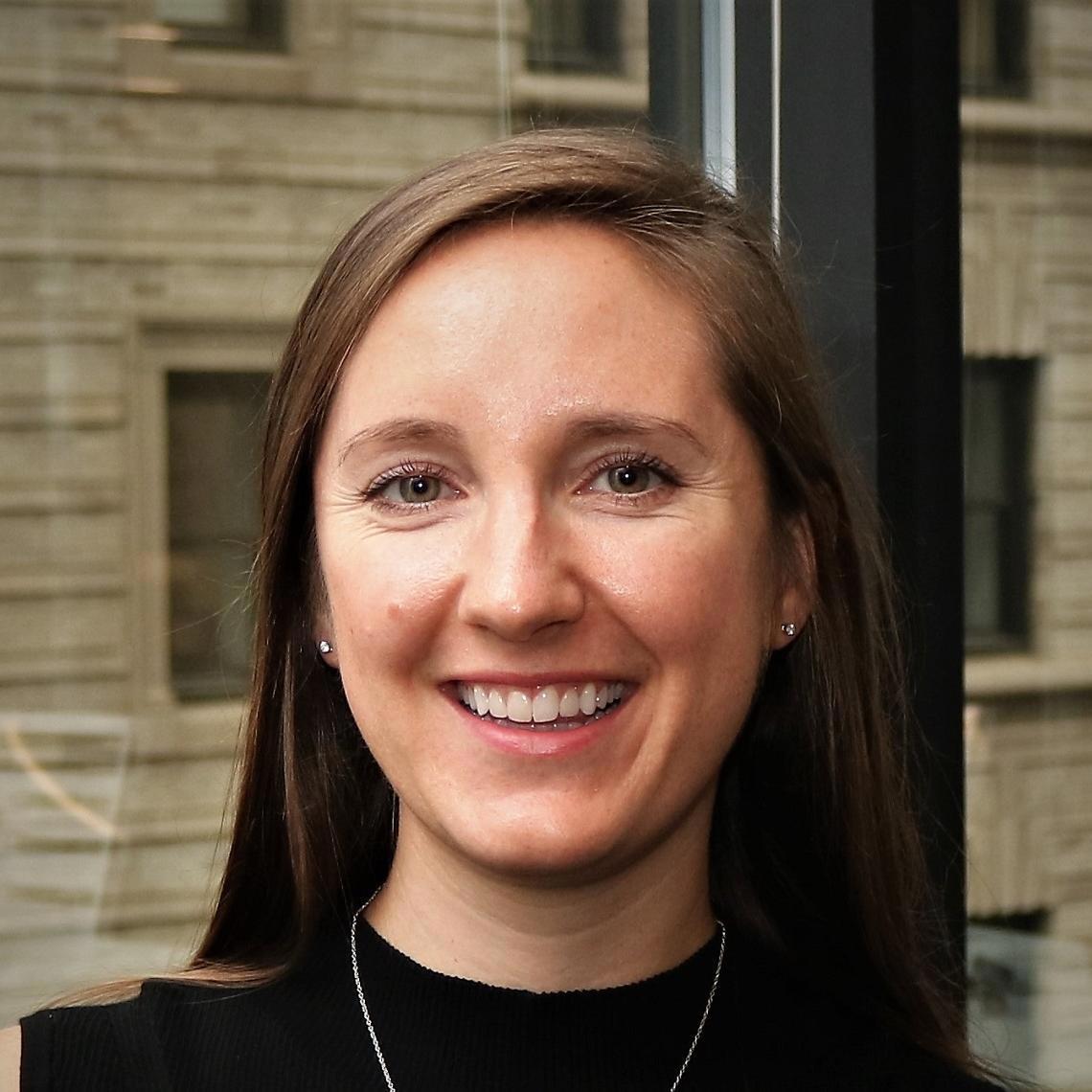 A woman with straight brown hair in a black shirt smiles in front of a window looking out onto a brown brick building.