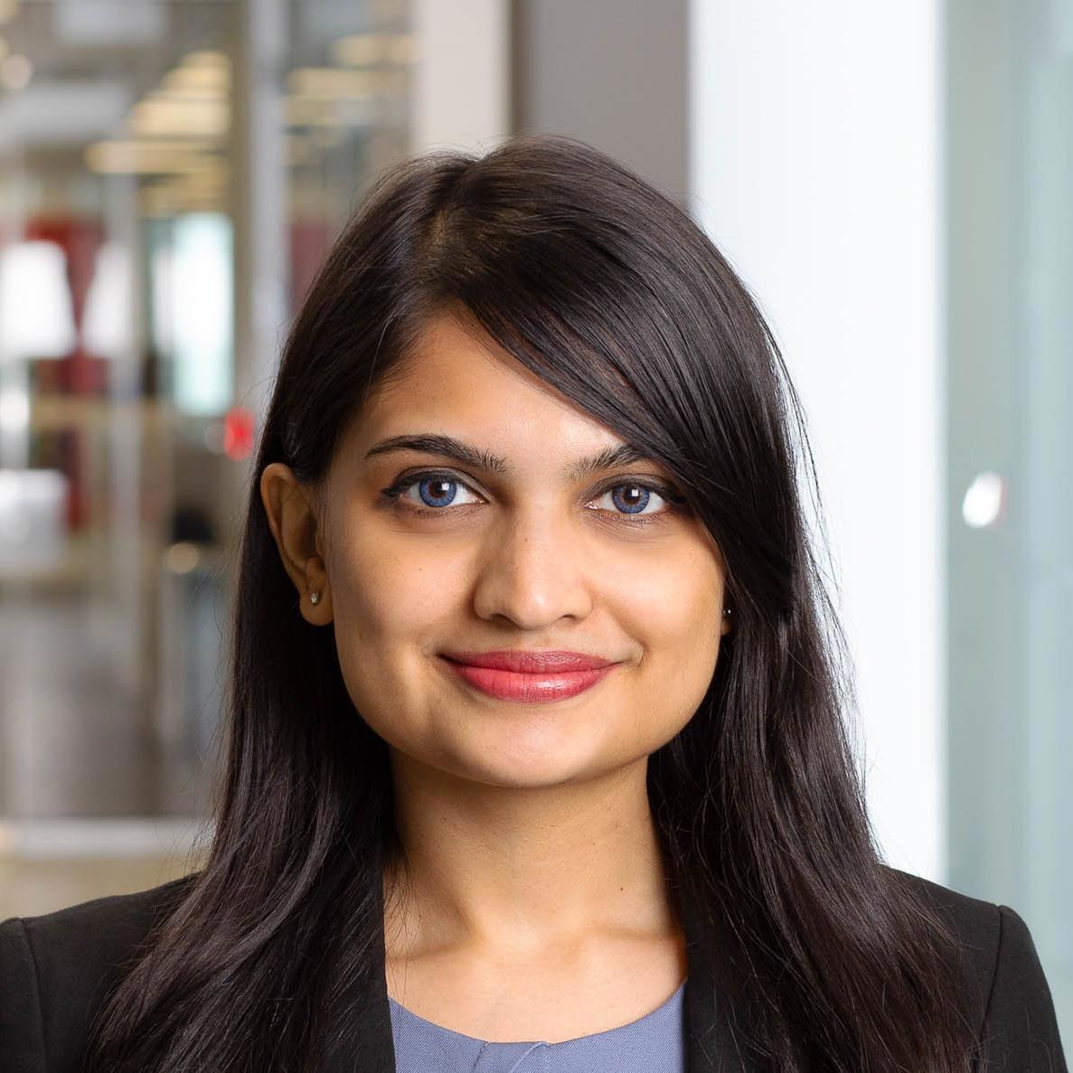 A close-up photo of a woman with long, straight black hair, smiling subtly at the camera. She is wearing a black blazer over a light blue top. The background is a blurred office setting with modern decor and soft lighting, which highlights her face.