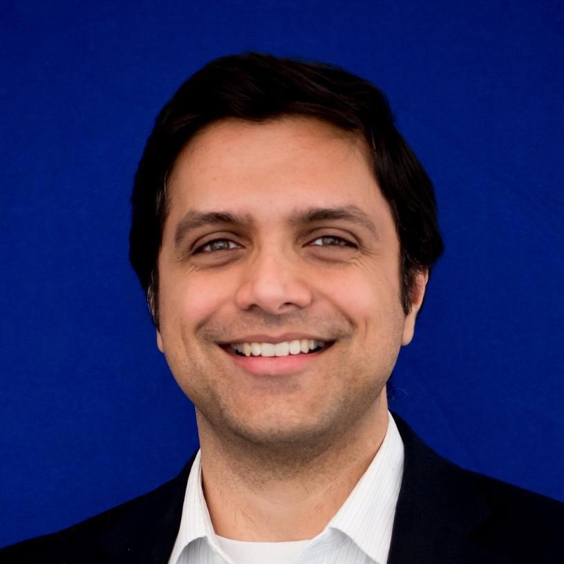 A close-up photo of a man with short black hair, smiling at the camera. He is wearing a dark suit jacket over a white collared shirt. The background is a solid bright blue, which contrasts with his dark hair and suit.