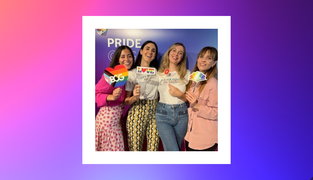 Four women smiling and holding colorful signs celebrating Pride are framed in a white square. The signs include a heart with the BCG logo in Pride colors, a "LOVE" sign with rainbow lettering, and a cloud with rainbow colors. They stand in front of a vibrant background with "PRIDE" prominently displayed. 