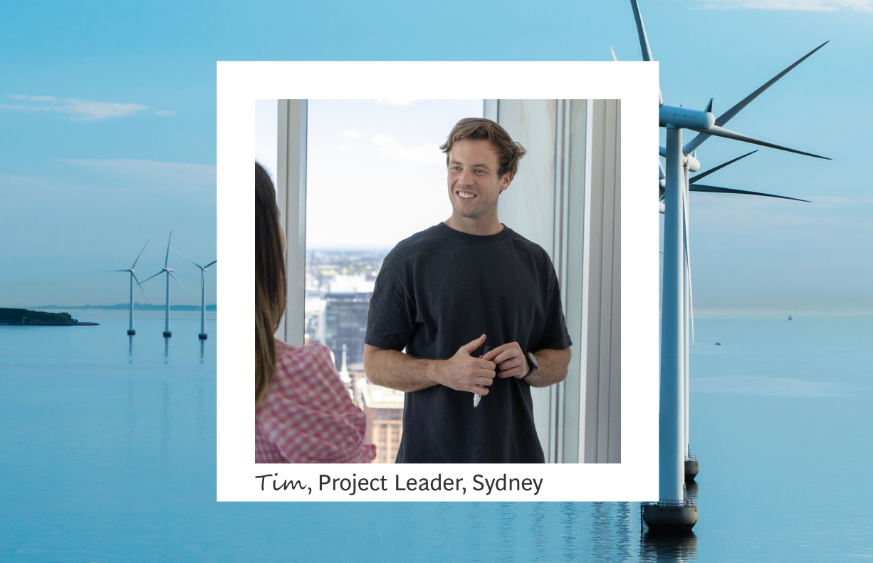 The image depicts Tim, a project leader in Sydney, engaged in a friendly conversation with a colleague inside a brightly lit office. The foreground shows Tim smiling and gesturing, wearing a casual black T-shirt, set against the backdrop of a cityscape visible through large windows set in a white square frame. In the background, an idyllic blue ocean with offshore wind turbines illustrates a commitment to sustainability, harmonizing with the serene, professional setting.