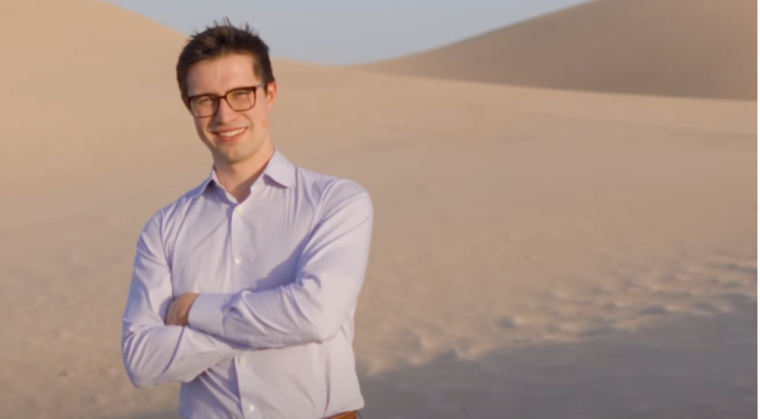 A man with short dark hair and glasses is standing with his arms crossed, wearing a light purple dress shirt. He is smiling and looking directly at the camera. The background features expansive sand dunes under a clear sky, suggesting a desert environment. The lighting indicates it is likely late afternoon or early morning, casting a soft glow over the scene.