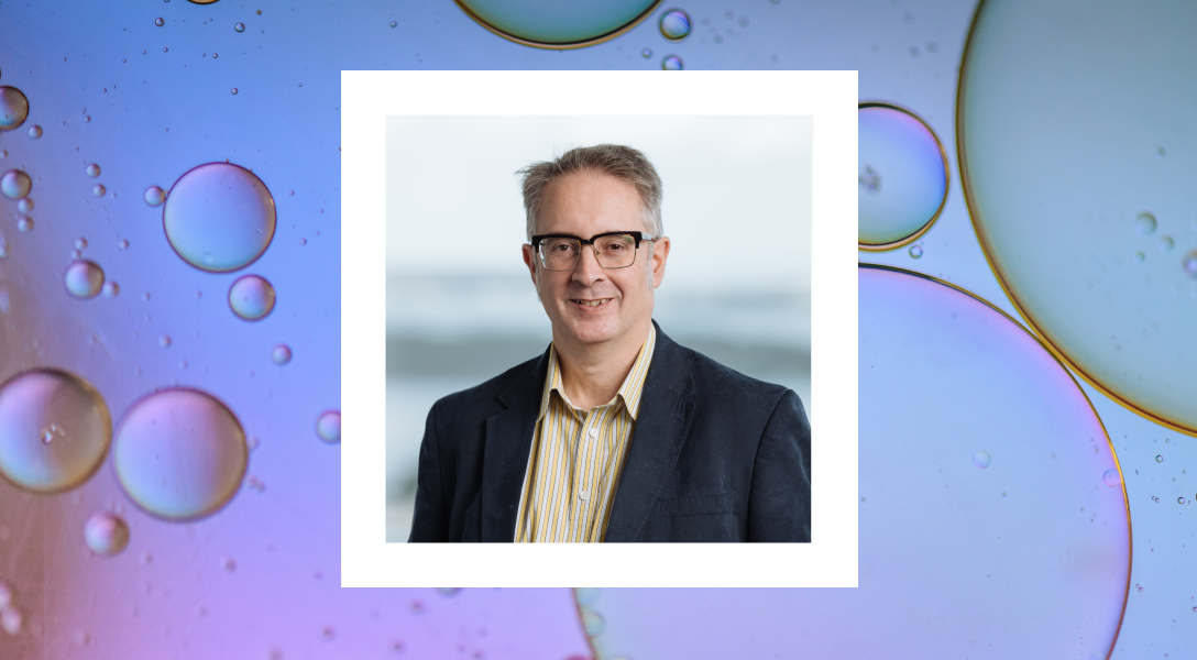 A close-up photo of a man with short gray hair and glasses, smiling at the camera. He is wearing a dark blazer and a striped shirt. The photo is framed in a polaroid-style white border. The background behind the polaroid features a colorful abstract design with large, translucent bubbles on a gradient of purple and blue.