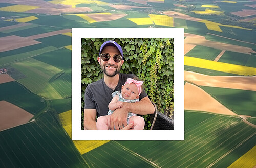 A smiling man wearing sunglasses and a cap, holding a baby girl dressed in a pink outfit with a matching bow headband. They are posing in front of a lush green vine-covered wall. The photo is framed within an aerial view of vast agricultural fields, showcasing varying shades of green and patterns, giving a picturesque countryside backdrop.