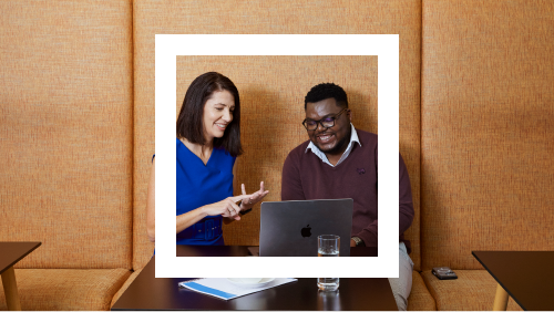 Two professionals, a woman in a blue blouse and a man in a maroon shirt, engaging in a discussion in front of a laptop on a wooden table, set against a textured orange wall.