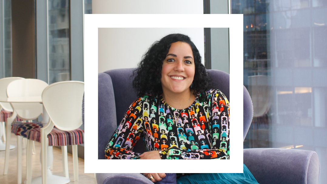 The image shows a woman sitting on a purple chair in a modern office environment with large windows in the background. She has curly, shoulder-length dark hair and is smiling warmly. She is wearing a colorful, patterned top featuring multiple stylized images of people. The photo is framed with a white square around her upper body, drawing attention to her smile and vibrant clothing. The background includes white tables and chairs with striped upholstery, adding to the contemporary feel of the space.
