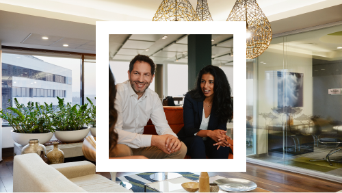 A photo of a man and a woman in business casual attire having a conversation in a well-appointed office lounge with plants and pendant lighting.