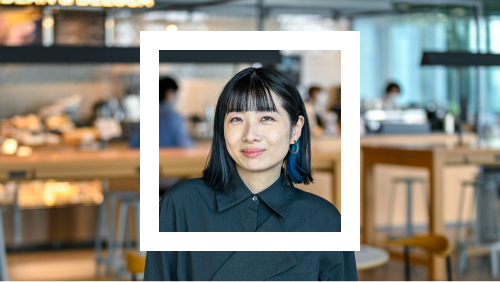 Portrait of a woman with shoulder-length black hair and bangs, wearing a dark blue shirt, set against a background of a spacious and well-lit cafe with wooden tables and chairs.