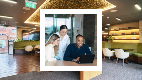 A photo of three colleagues looking at a laptop together, set against a cozy office break area with bookshelves and modern furniture