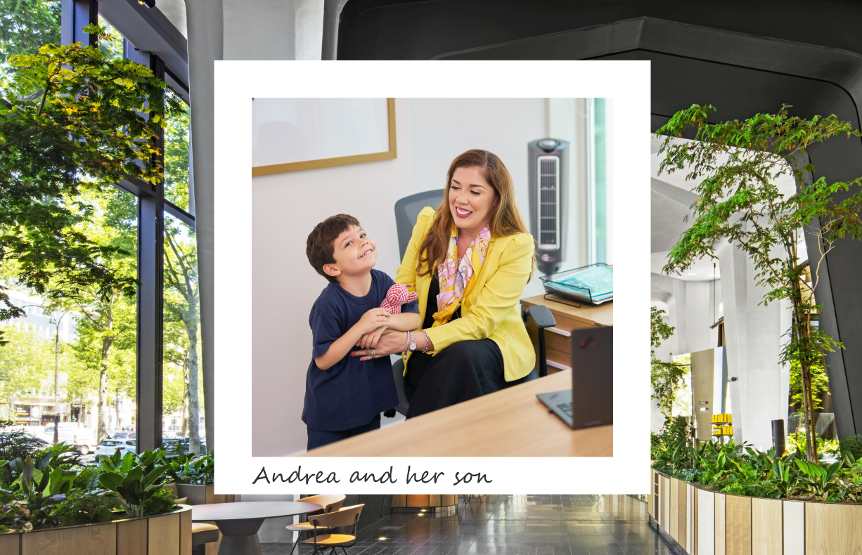 A joyful moment captured between Andrea and her son. Andrea, dressed in a bright yellow blazer and patterned scarf, is seated at her desk in a modern office setting, smiling warmly at her young son, who is standing beside her in a navy blue shirt, looking up at her with a wide smile. This heartwarming image is framed within a larger background of a spacious, green-filled office atrium with large windows, bringing in ample natural light and highlighting the connection between work and family life. The caption reads, "Andrea and her son."