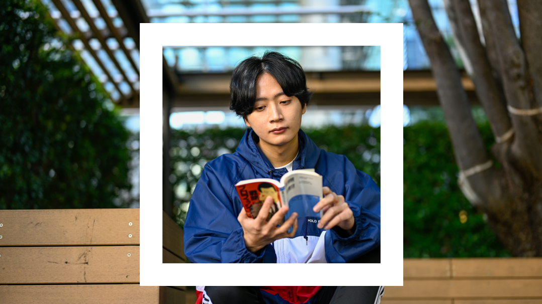 The image captures a young man deeply engrossed in reading a book while sitting on a park bench. He is dressed casually in a blue jacket and a red shirt visible underneath. The scene is framed within a white border, highlighting him against a backdrop of lush greenery and modern architectural elements. 