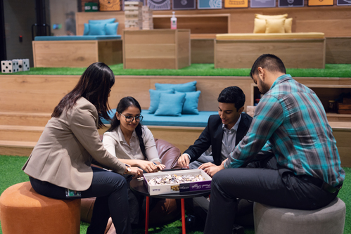Two professionals, a woman in a blue blouse and a man in a maroon shirt, engaging in a discussion in front of a laptop on a wooden table, set against a textured orange wall.