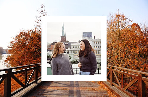 Two women engaged in a conversation on a roof deck. One woman, with blonde hair, looks intently at the other, who has dark hair, as they both smile. In the background, there's a view of a cityscape featuring a prominent church spire. The photo is framed within a scene of autumn, with a wooden bridge and trees showcasing vibrant orange foliage, providing a warm and serene ambiance.