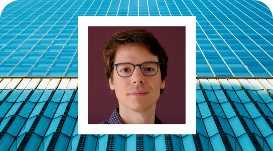 Joerg Schneider, Principal AI engineer in Munich, wearing specs and facing towards towards the camera smiling, his photo is inside a white bordered frame set against a background of blue building windows 