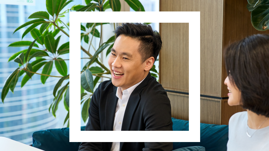 This image captures a joyful interaction in a modern office setting, featuring a young Asian man, dressed in a black blazer and white shirt, framed within a white border. He is smiling broadly while conversing with a colleague, depicted partially to the right. The background includes lush indoor plants and a view of an urban skyline through large windows, contributing to a bright and vibrant workspace. 