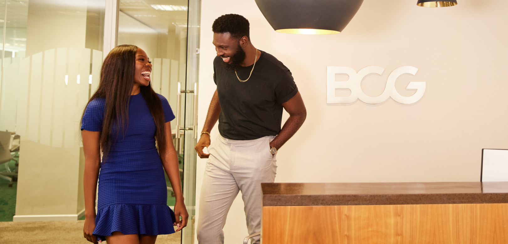 Two individuals are standing in an office lobby. The person on the left is wearing a blue dress and the person on the right is wearing a black t-shirt and white pants. They appear to be conversing or walking together. In the background, there’s a reception desk with the logo ‘BCG’ prominently displayed above it, indicating this may be the office of Boston Consulting Group 