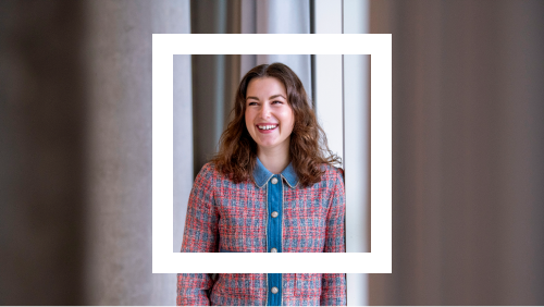 Portrait of a cheerful young woman with wavy brown hair, wearing a colorful checkered blazer, set against a soft-focused architectural background.