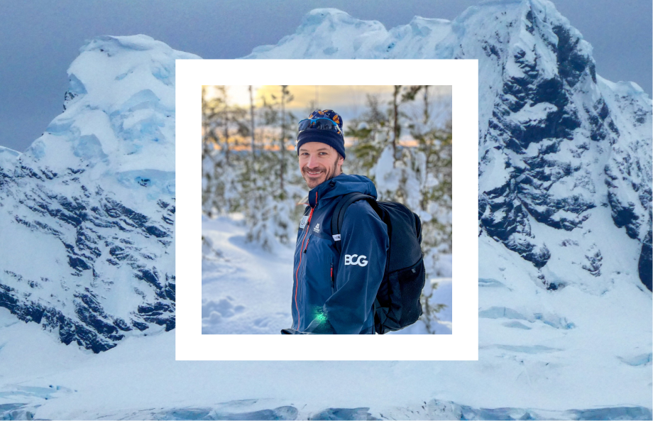 This image depicts a hand holding a photograph with a snowy mountain backdrop, featuring icy peaks and rugged terrain. Inside the photograph, a man is pictured in a winter setting, surrounded by snow-covered trees. He is wearing a warm hat, a jacket with the logo "BCG," and is carrying a backpack, smiling at the camera. 