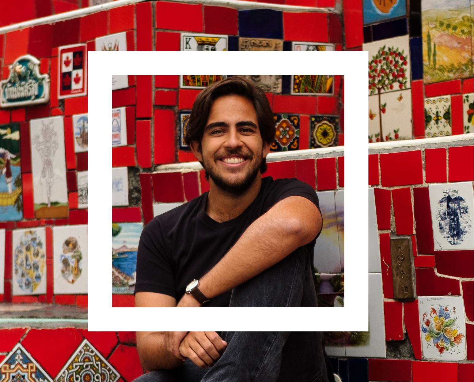 A man is sitting in front of a vibrant mosaic wall composed of variously patterned tiles. The individual is wearing a black t-shirt and has one arm resting on a raised knee. The mosaic features an array of colors and designs, including floral patterns, geometric shapes, and artistic representations that contribute to the wall's eclectic aesthetic. 