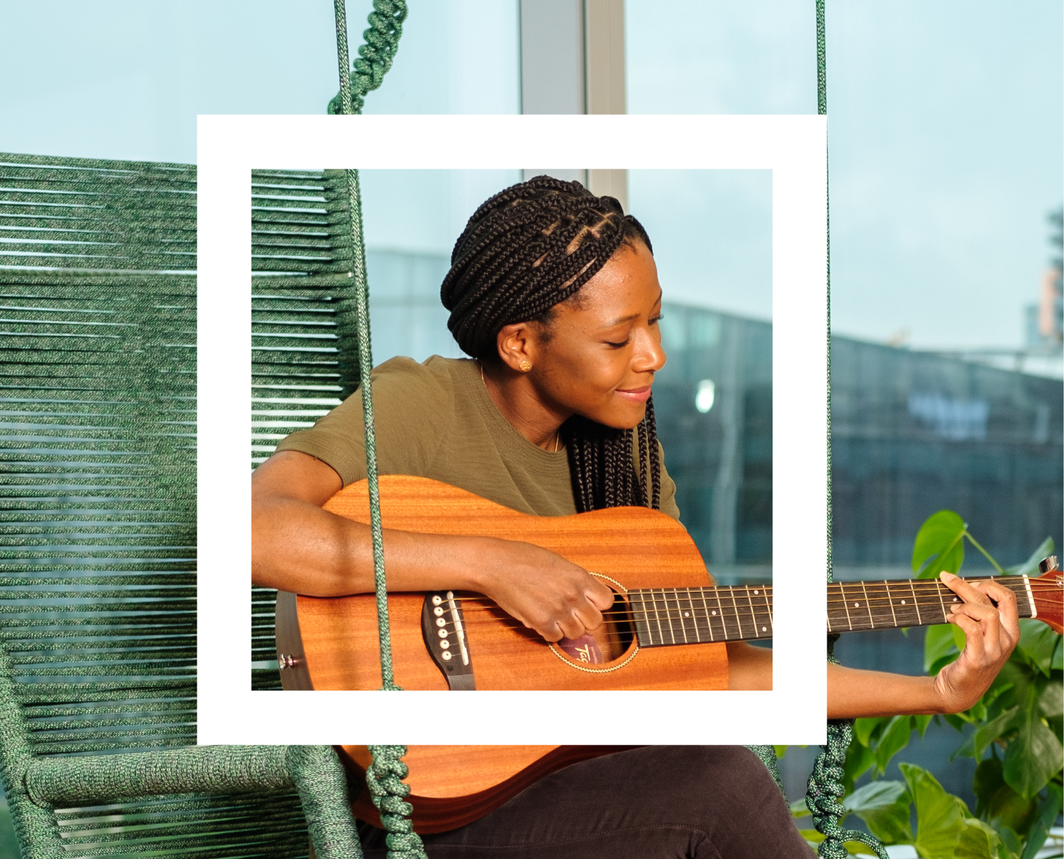 A woman with braided hair is seated playing an acoustic guitar. The setting appears to be indoors with a large window in the background, suggesting a relaxed and casual atmosphere. Visible elements include the guitar’s sound hole, strings, and fretboard as the person’s fingers are positioned to play a chord. Green blinds partially cover the window, complementing the serene ambiance. 