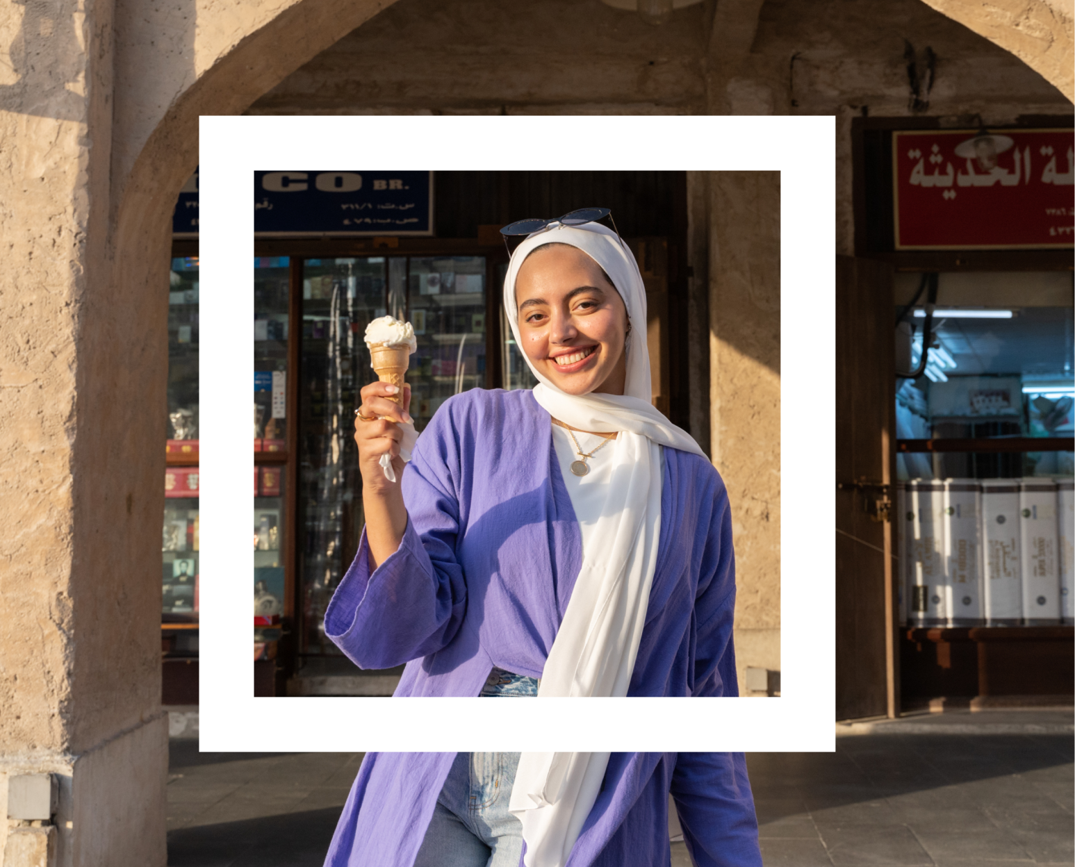 A Polaroid-style photograph features a young woman smiling and holding an ice cream cone. She is wearing a white hijab, a light purple cardigan, and blue jeans. She also has sunglasses perched on her head and is accessorized with a gold necklace. The setting appears to be an outdoor market or a traditional area with stone arches and storefronts in the background, bathed in warm sunlight.