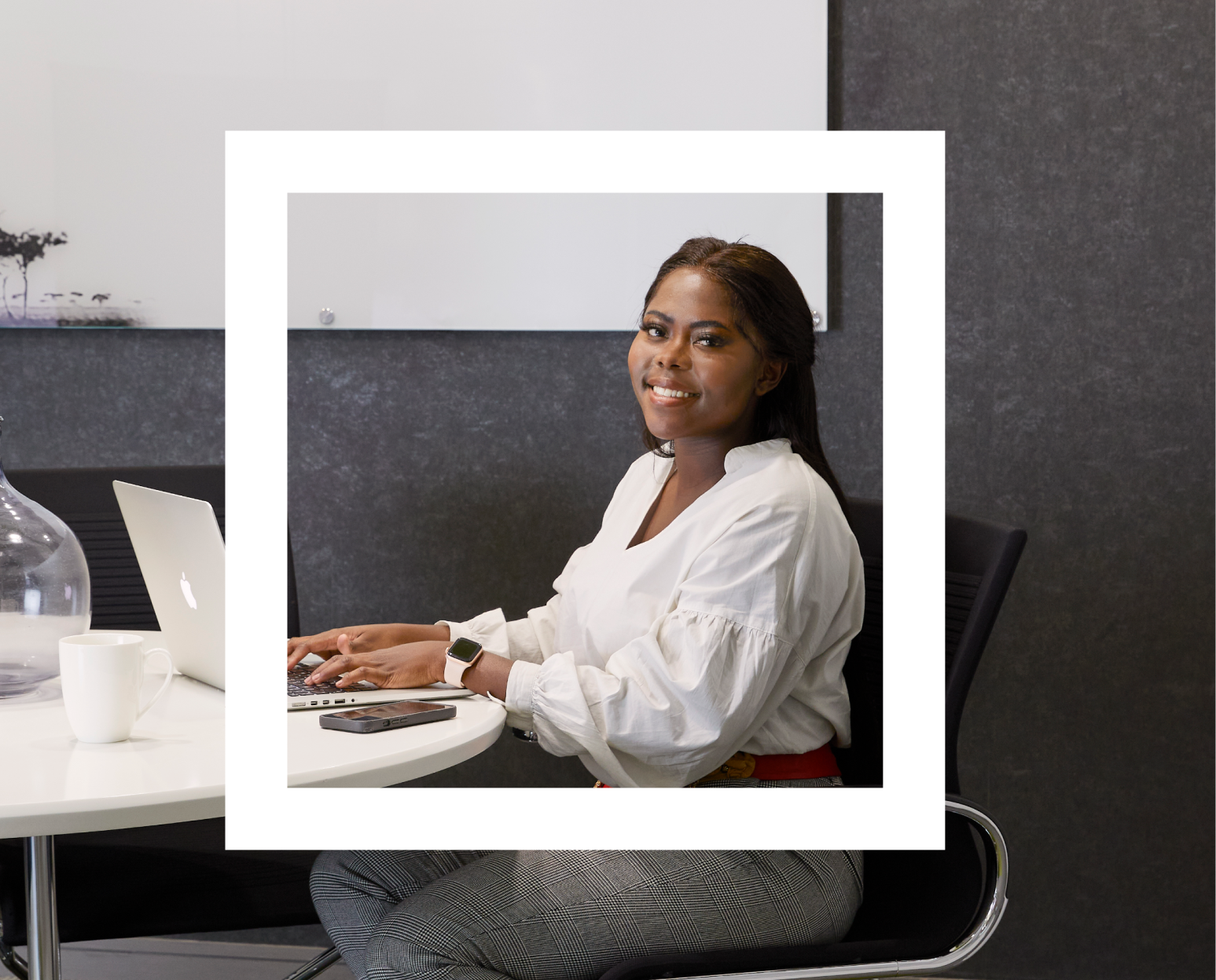 A Polaroid-style photograph features a young woman sitting at a white round table in a modern office setting. She is smiling and looking at the camera while typing on a laptop. She is wearing a white blouse and checkered pants, with a smartwatch on her wrist. On the table in front of her are a white mug, a smartphone, and a glass vase. The background includes a dark wall and a whiteboard, creating a professional and sleek work environment.
