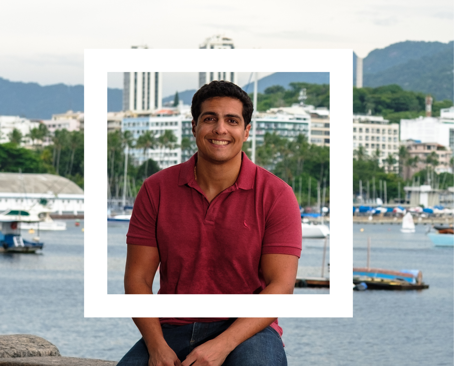 A man in a red polo shirt is sitting on a stone ledge with a scenic waterfront and buildings in the background. The setting includes mountains and a marina with boats docked. 