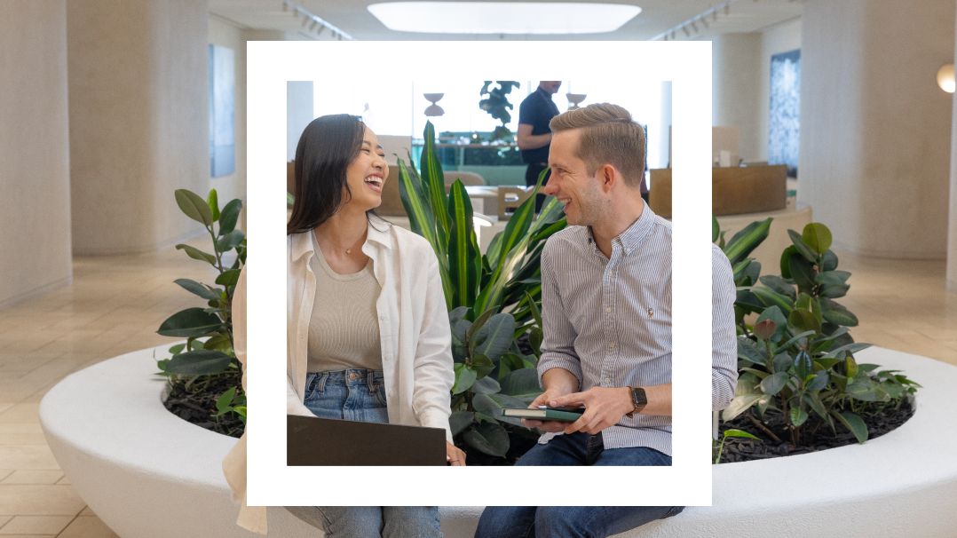 A framed image of a young Asian woman and a Caucasian man seated on a white circular bench surrounded by lush green plants in a bright, modern office space. They are both smiling and engaging in a lively conversation, with the man holding a tablet and the woman a laptop. 