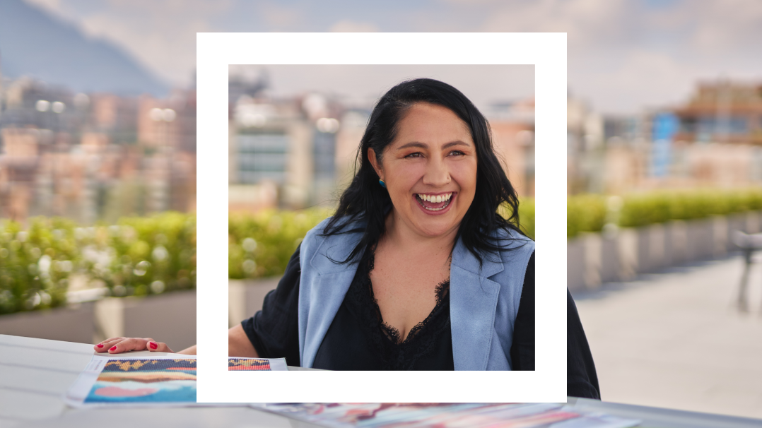 The image shows a woman laughing joyfully, with long dark hair, wearing a light blue sleeveless blazer over a black shirt. She is sitting outdoors at a table with a blurred cityscape and greenery in the background. The image has a white frame or border overlaid, creating a layered effect that highlights her expression and adds depth to the composition. Her cheerful demeanor and casual yet professional attire suggest a moment of relaxed conversation or enjoyment.