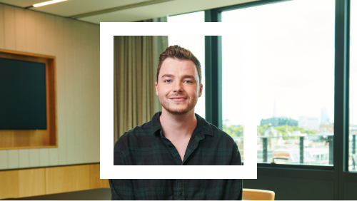 Portrait of a man with short brown hair, wearing a dark green checkered shirt, standing in a modern office room with large windows showcasing a city view in the background.
