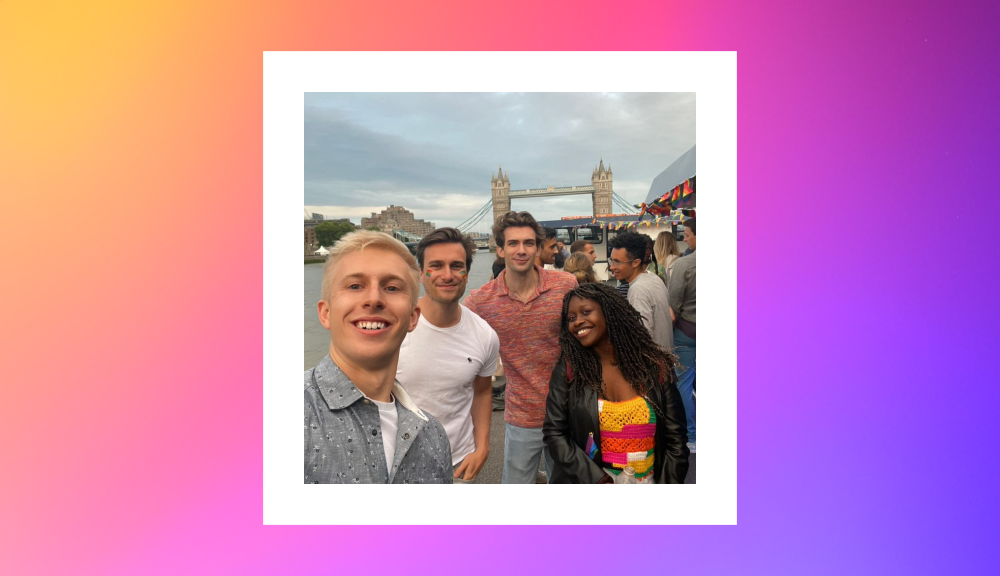 Four friends smiling and posing for a selfie with the iconic Tower Bridge in London in the background. The group includes two men in casual shirts, another man in a colorful patterned shirt, and a woman wearing a vibrant rainbow-striped top and jacket. The scene captures a joyful and inclusive atmosphere, with a festive crowd and Pride decorations adding to the celebratory mood.