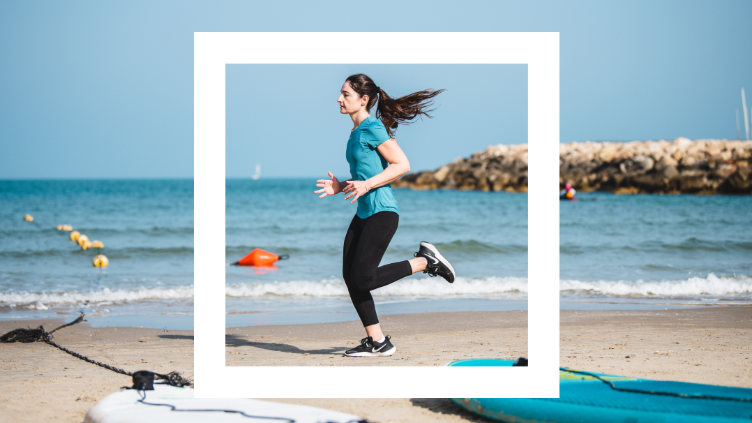 The image captures a dynamic scene of a woman running along a sandy beach, framed within a white border. She is dressed in athletic wear, consisting of a teal t-shirt and black leggings, and her hair flows behind her as she moves. The background features a calm sea and a rocky breakwater under a clear blue sky.
