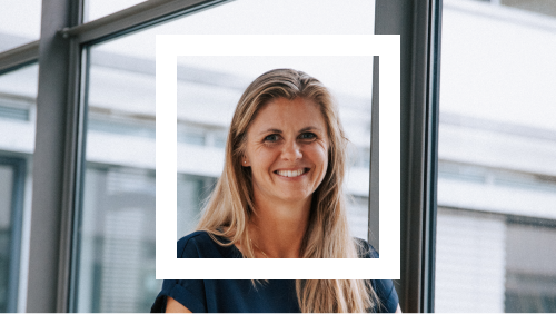 Portrait of a smiling woman with long blonde hair, wearing a dark blouse, posed in front of large glass windows in a well-lit office setting.