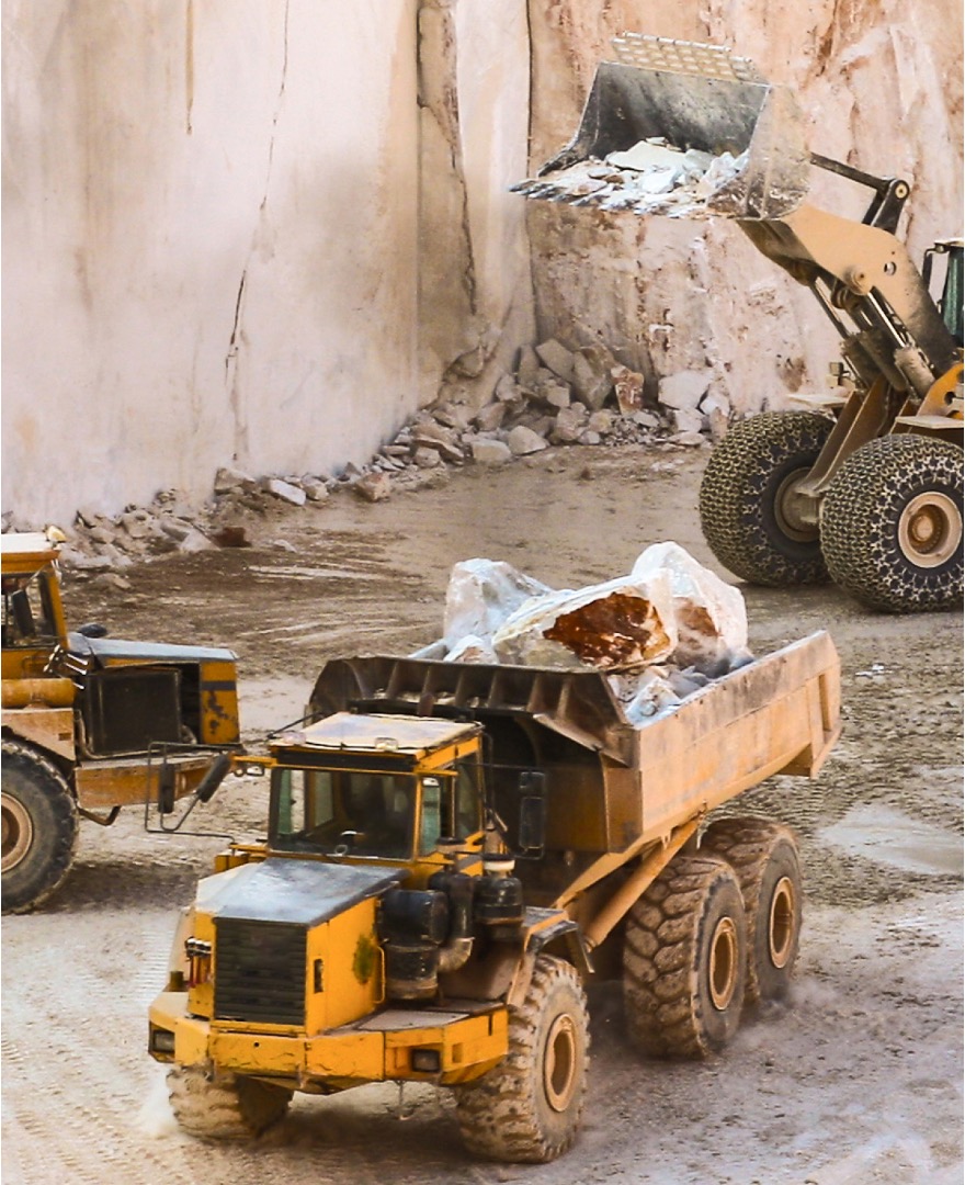 A large yellow dump truck is being loaded with rocks by an excavator in a quarry. The truck is positioned in the foreground with its bed raised, and chunks of stone are visible inside it. In the background, a high wall of exposed earth indicates an active excavation site. Dust and tire tracks suggest recent heavy machinery movement
