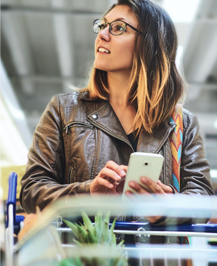 A woman stands behind a shopping cart holding a smartphone. The individual is wearing a brown leather jacket and has shoulder-length hair. The setting appears to be indoors, possibly a supermarket or store, indicated by the blurred background suggesting aisles and merchandise shelves 