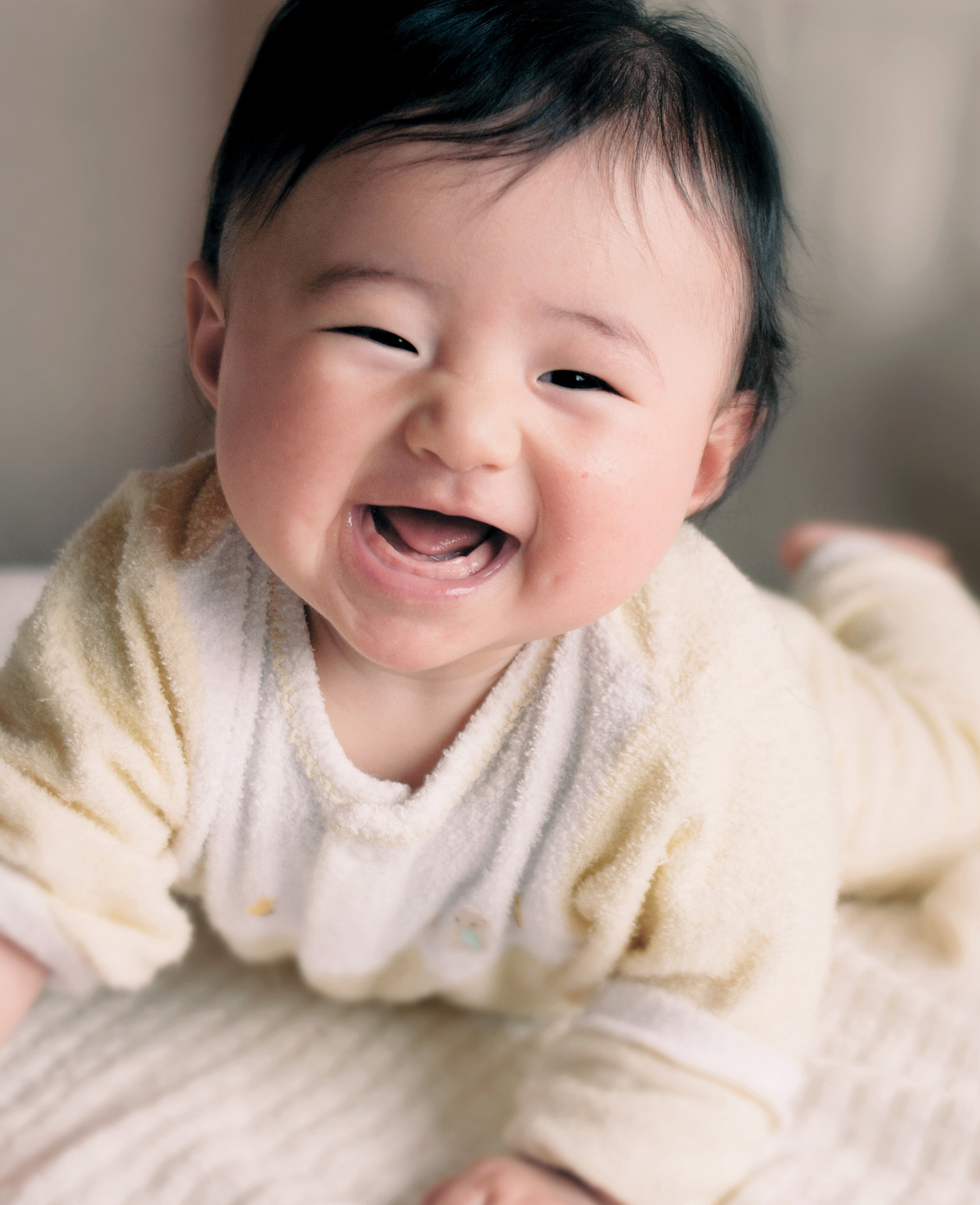 A close-up image of a baby wearing a white outfit, laughing and crawling.The background is soft and indistinct, suggesting an indoor setting with natural lighting. 