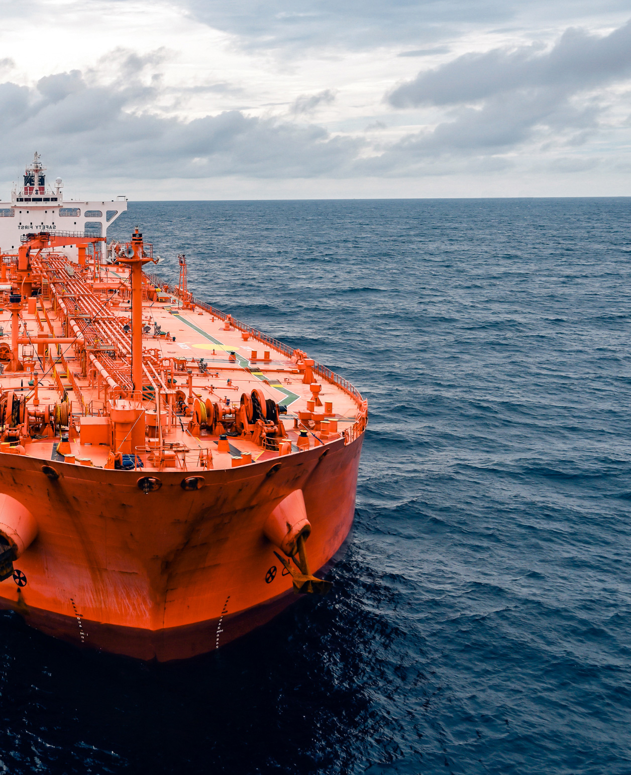 A large orange-hulled tanker ship at sea, with a cloudy sky above and calm blue water surrounding it. The deck of the ship is equipped with various mechanical structures and equipment, indicating its industrial purpose. The perspective is from the front of the vessel looking towards the back, capturing the vastness of the ocean and the scale of maritime transport. 