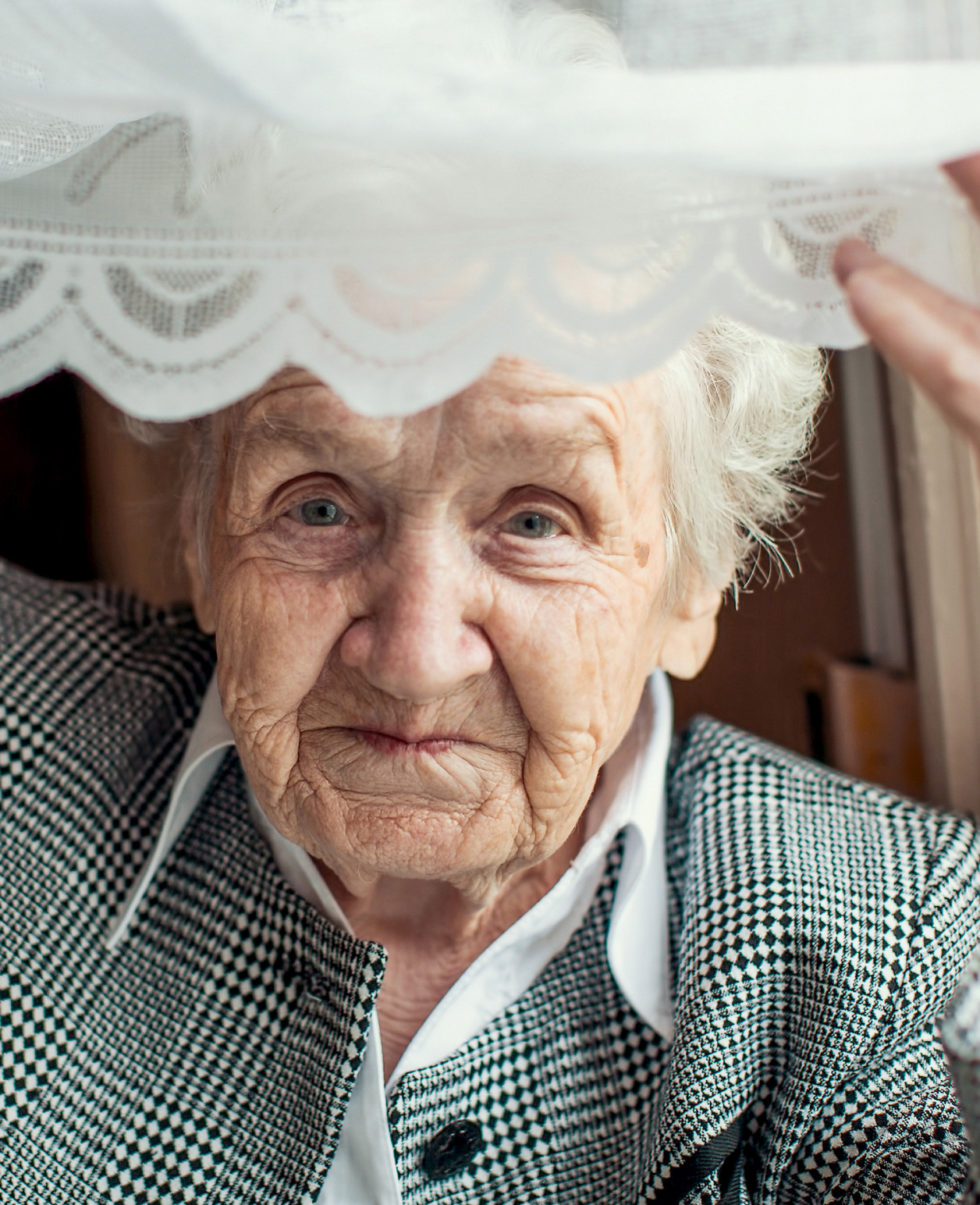 Close-up of an elderly person with wrinkled skin, looking directly at the camera. The individual is wearing a houndstooth patterned jacket over a white shirt and has white hair partially visible under a lace-trimmed garment being held up with one hand 