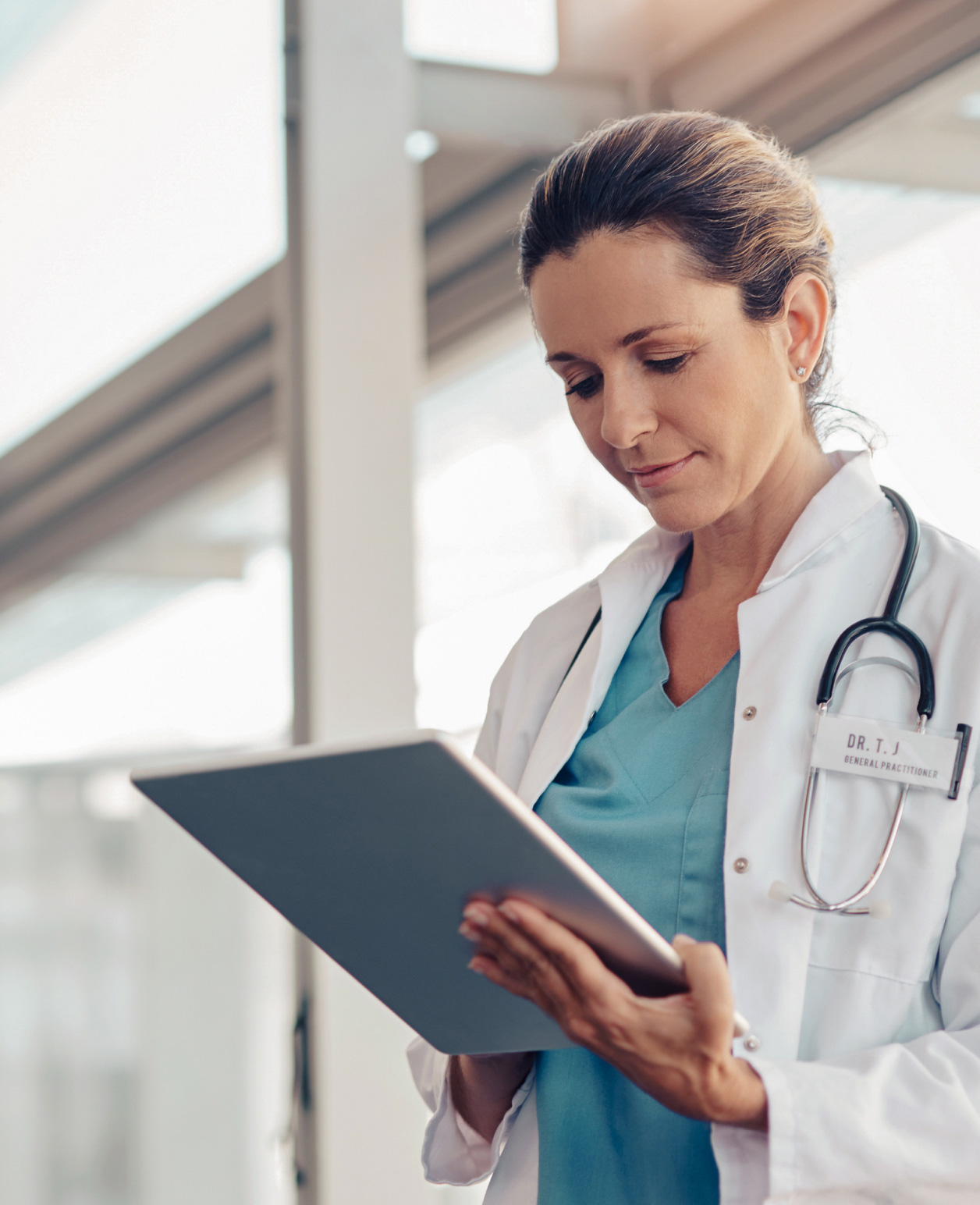 A person in a white lab coat, identified as a doctor by the badge and stethoscope, is holding and looking at a clipboard. The background suggests an indoor setting with natural light coming through windows 