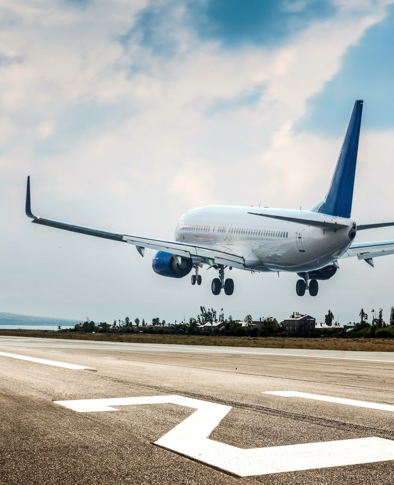 A commercial airplane with blue and white livery is captured in the midst of landing, with its landing gear extended towards a runway. The perspective is from ground level, looking up at the plane as it descends against a backdrop of a partly cloudy sky. In the foreground, runway markings in the form of large white arrows point towards the direction of the landing aircraft, suggesting guidance for pilots during takeoff and landing procedures. 