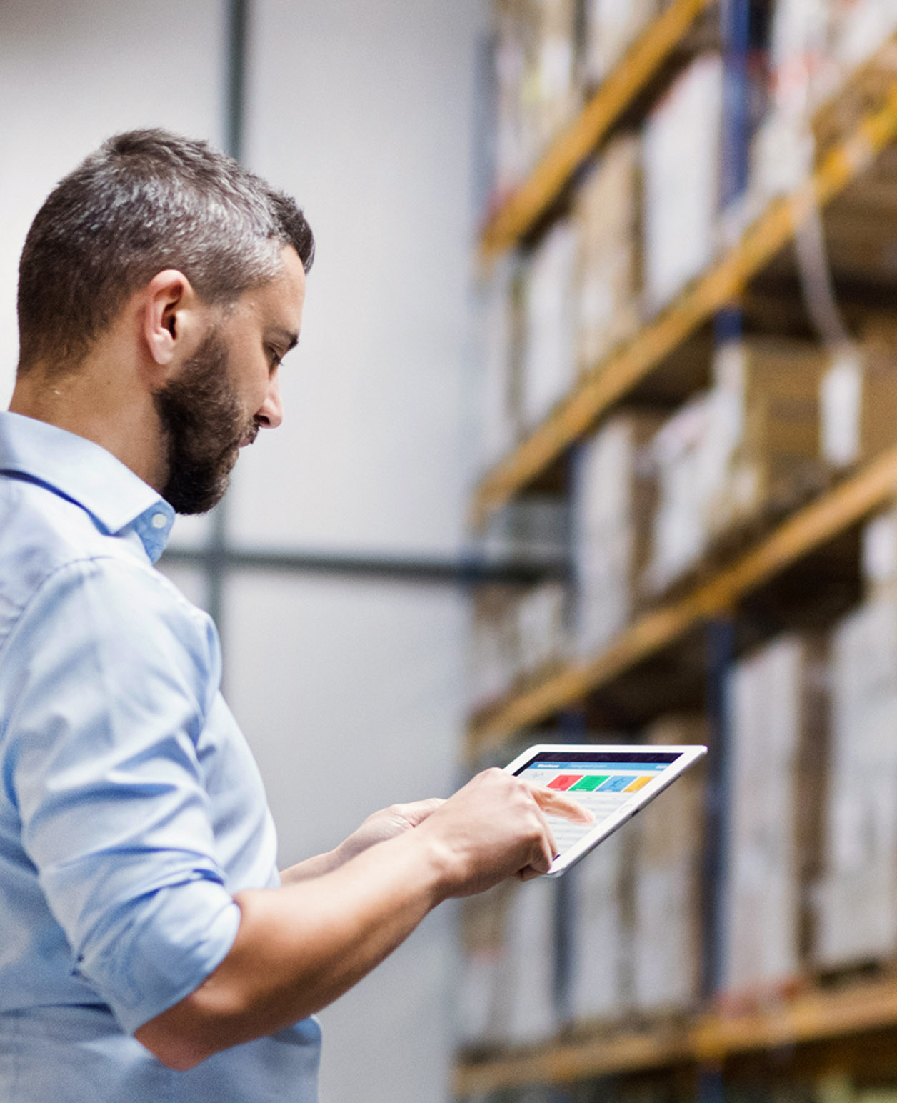 A person is standing in a warehouse, holding a tablet displaying colorful bar graphs. The individual is wearing a light blue shirt and appears to be interacting with the data on the screen, possibly managing inventory or reviewing performance metrics. The background shows shelves stocked with various items in an organized manner, indicative of an operational storage facility.
